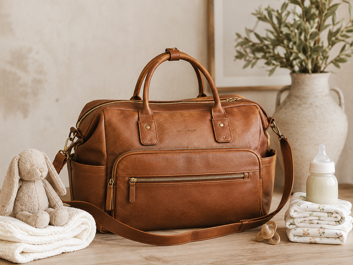 Sac à langer en cuir marron posé sur une table en bois, entouré d'un doudou lapin, d'un biberon, d'une tétine et de langes pliés